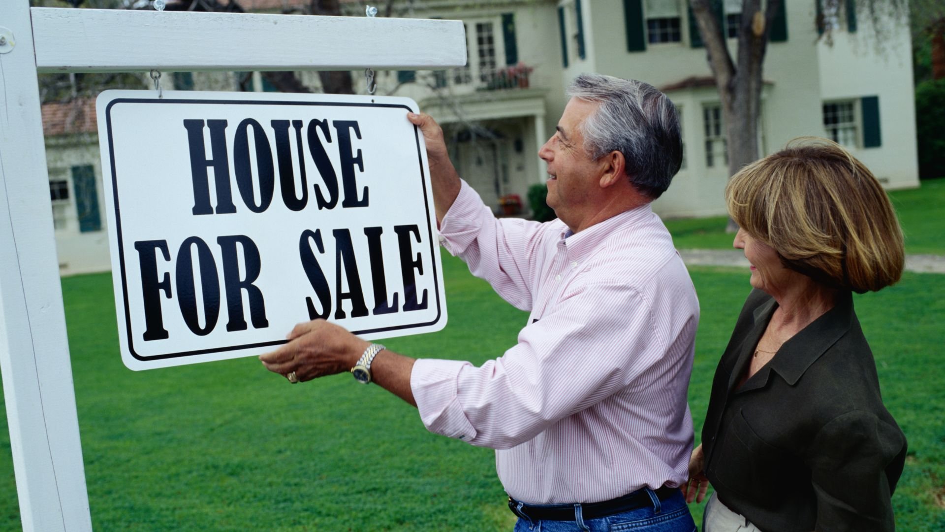 Older couple placing House For Sale sign in front of suburban home